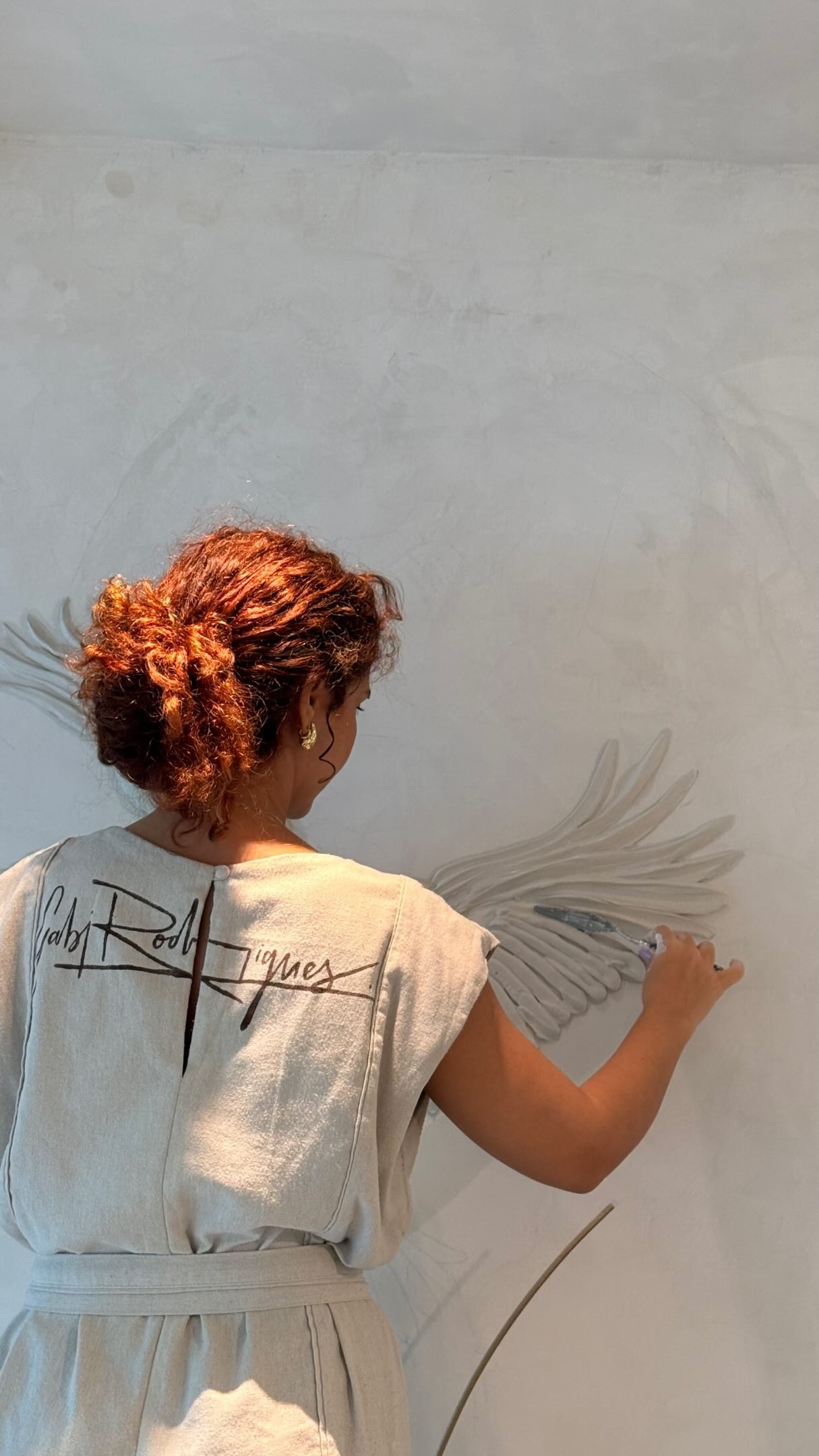 Gabi Rodrigues, de costas, pintando um mural de asas em relevo branco, com a própria assinatura estampada no uniforme
